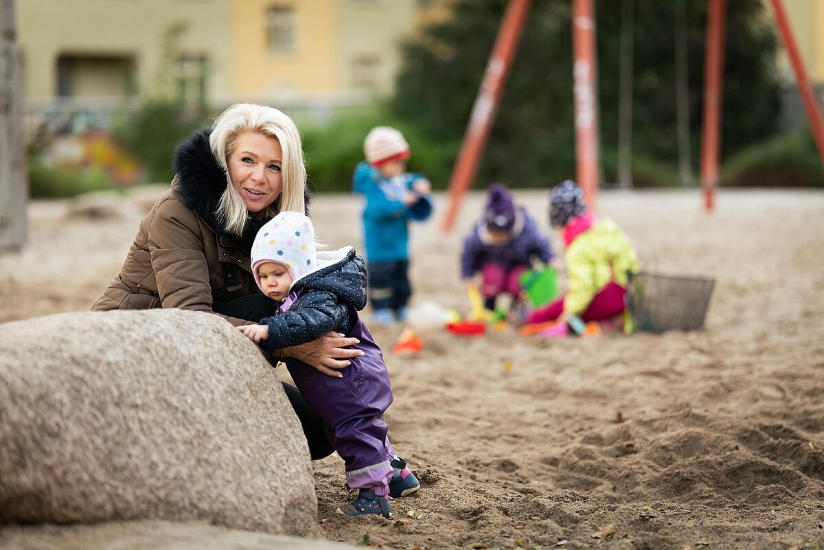 Kinder und Tagesmutter in einer Sandkisten sitzend vor einem großen Stein 
