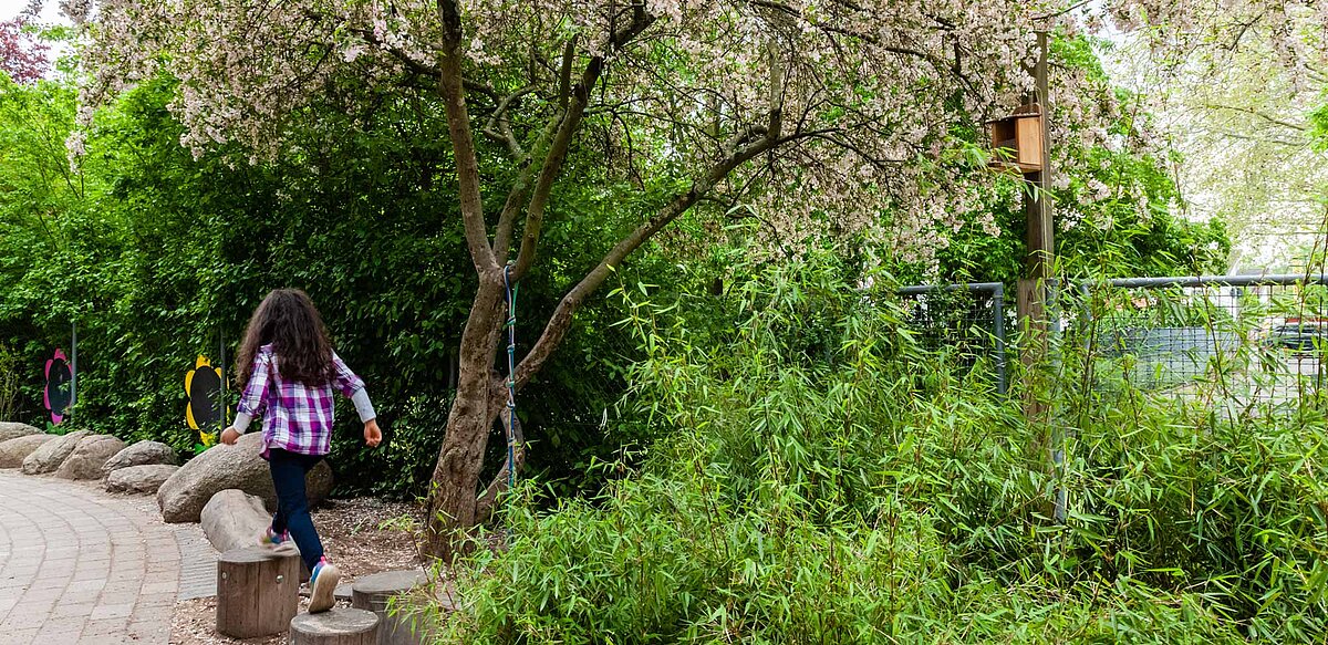 Aussenaufnahme, viel grüne Hecken. Ein Kind läuft am linken Rand