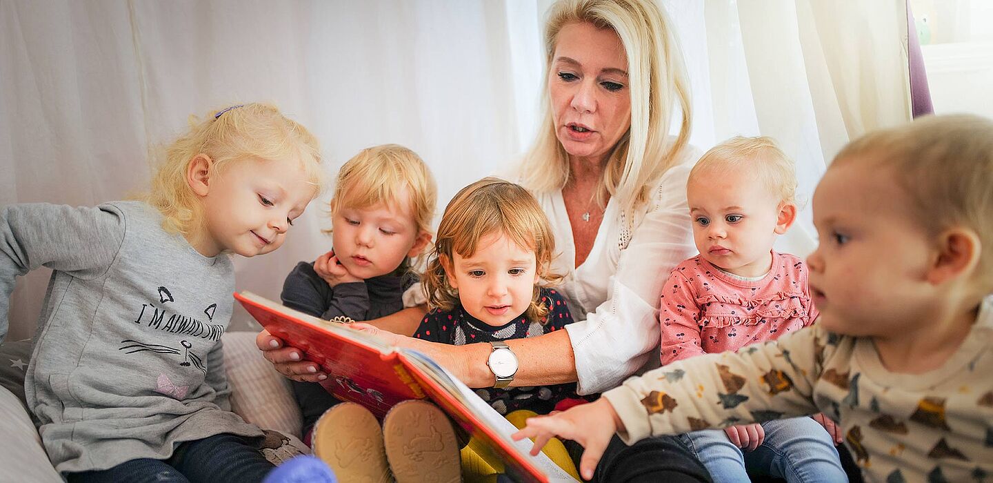 Tagesmutter sitzt mit mehreren Kindern auf dem Boden und schaut ein Buch an die Kinder sitzen drum herum